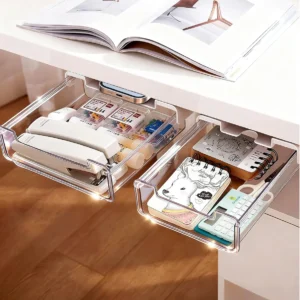 Dos cajones organizadores transparentes debajo de un escritorio blanco, llenos de suministros de oficina como grapadoras, notas y calculadora.