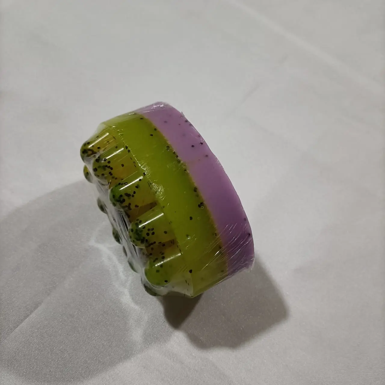 Jabón masajeador en capas verde y morado con semillas, envuelto en plástico.