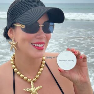 Mujer sonríe en la playa sosteniendo producto Marbella Beauty Matificante Facial, luce sombrero, gafas, collar y aretes dorados de estrella.