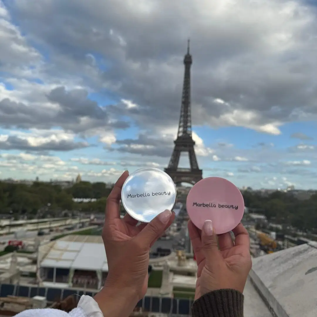 Dos compactos de matificante facial Marbella Beauty, uno transparente con reflejo del cielo parisino y otro rosa, Eiffel de fondo.