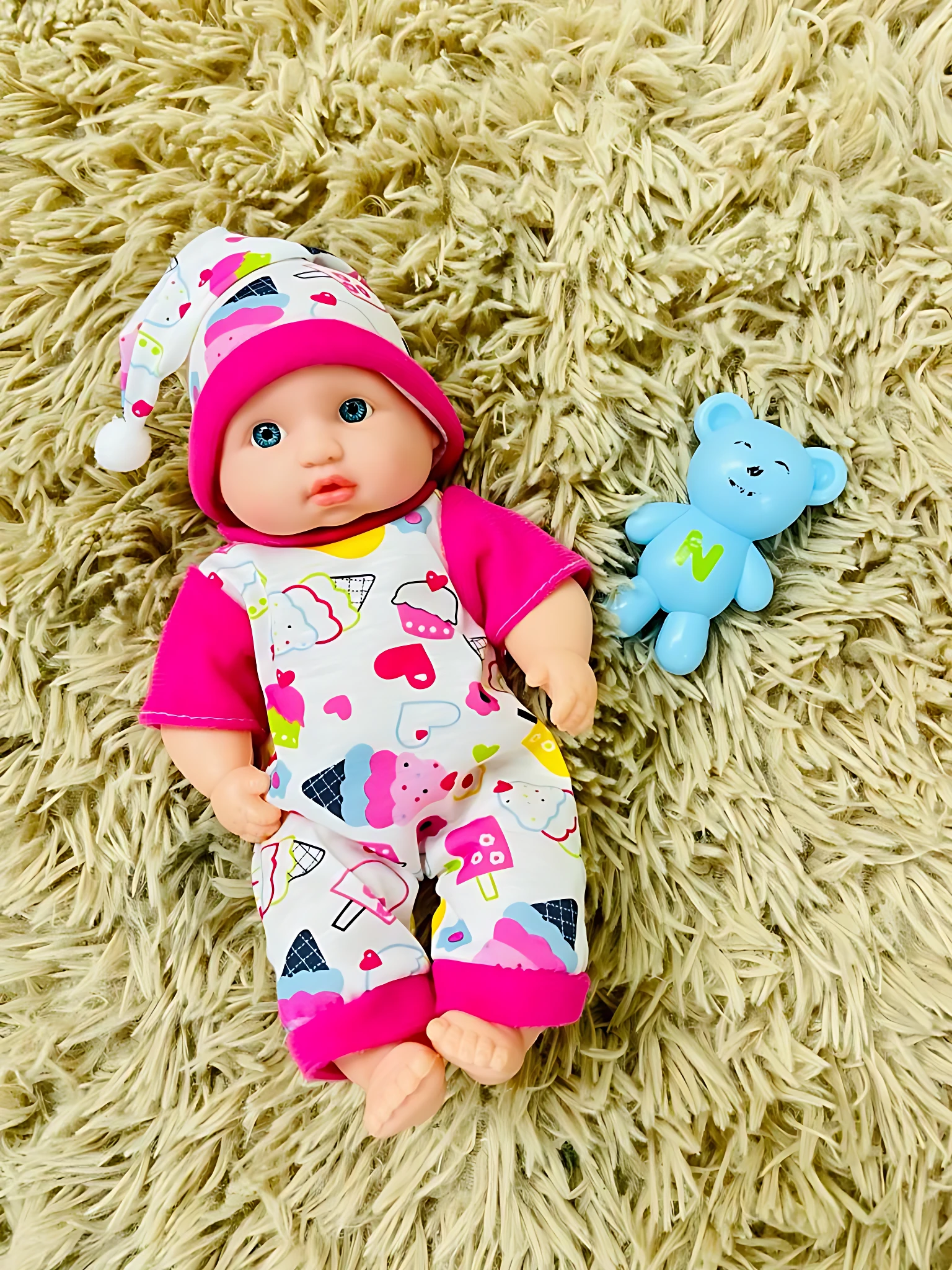 Muñeco bebé con pijama de helados y gorro, junto a un osito de plástico azul.