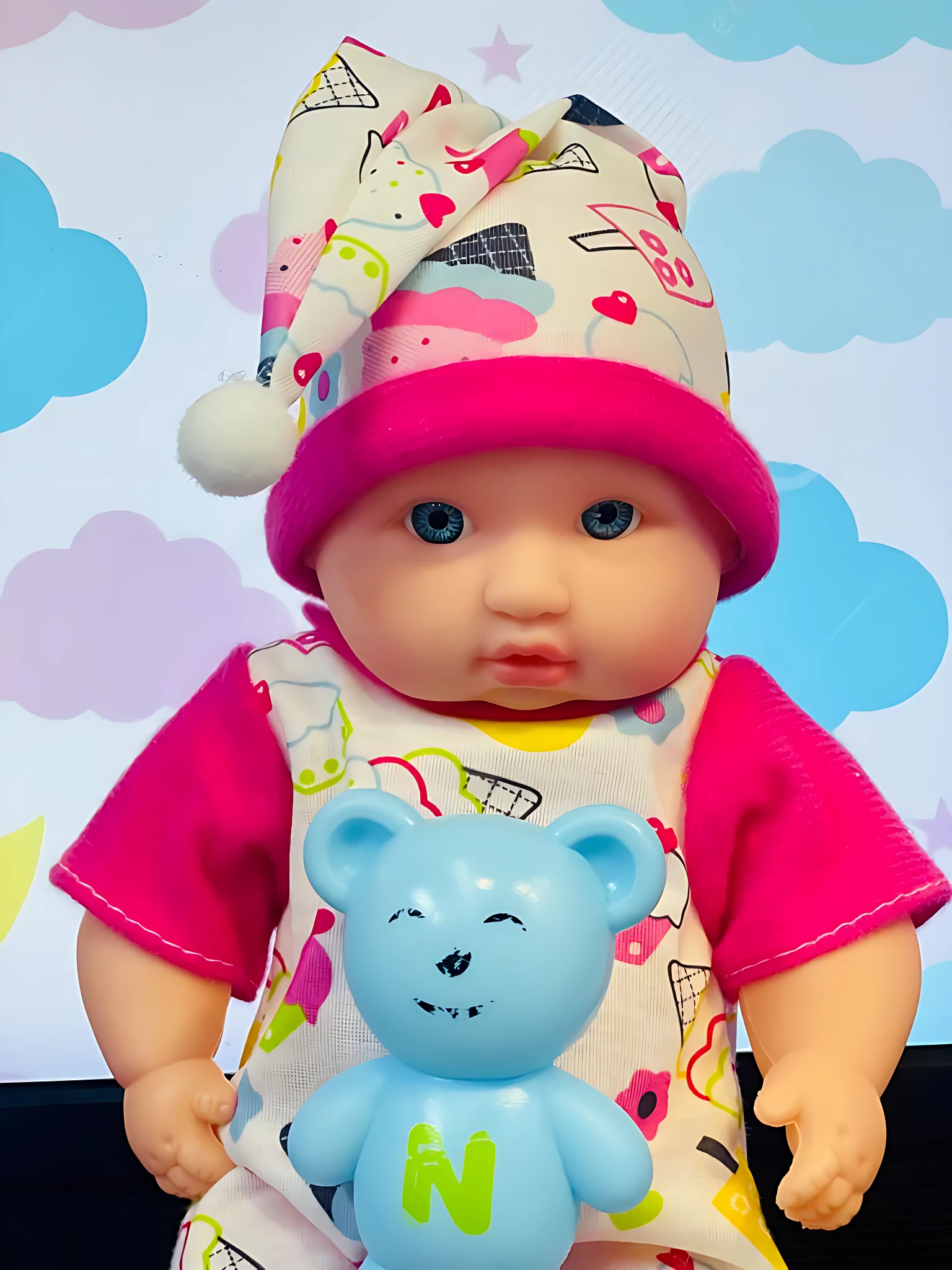 Bebé muñeco con pijama estampado de nubes y corazón, gorro de dormir con pompón, y osito azul de plástico con letra N.