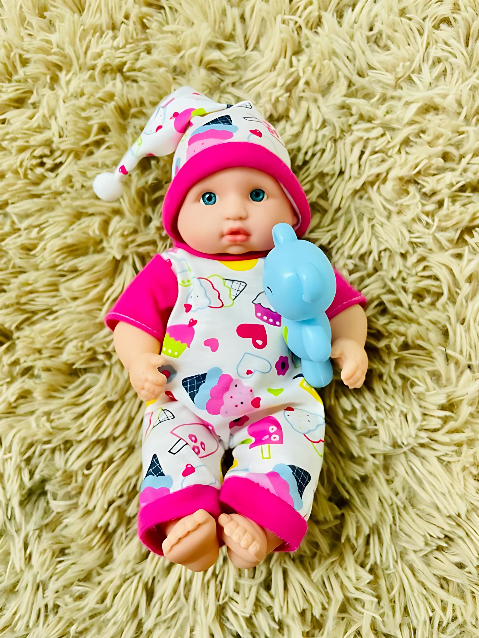 Muñeco bebé con pijama de cupcakes y osito de plástico azul sobre alfombra peluda.