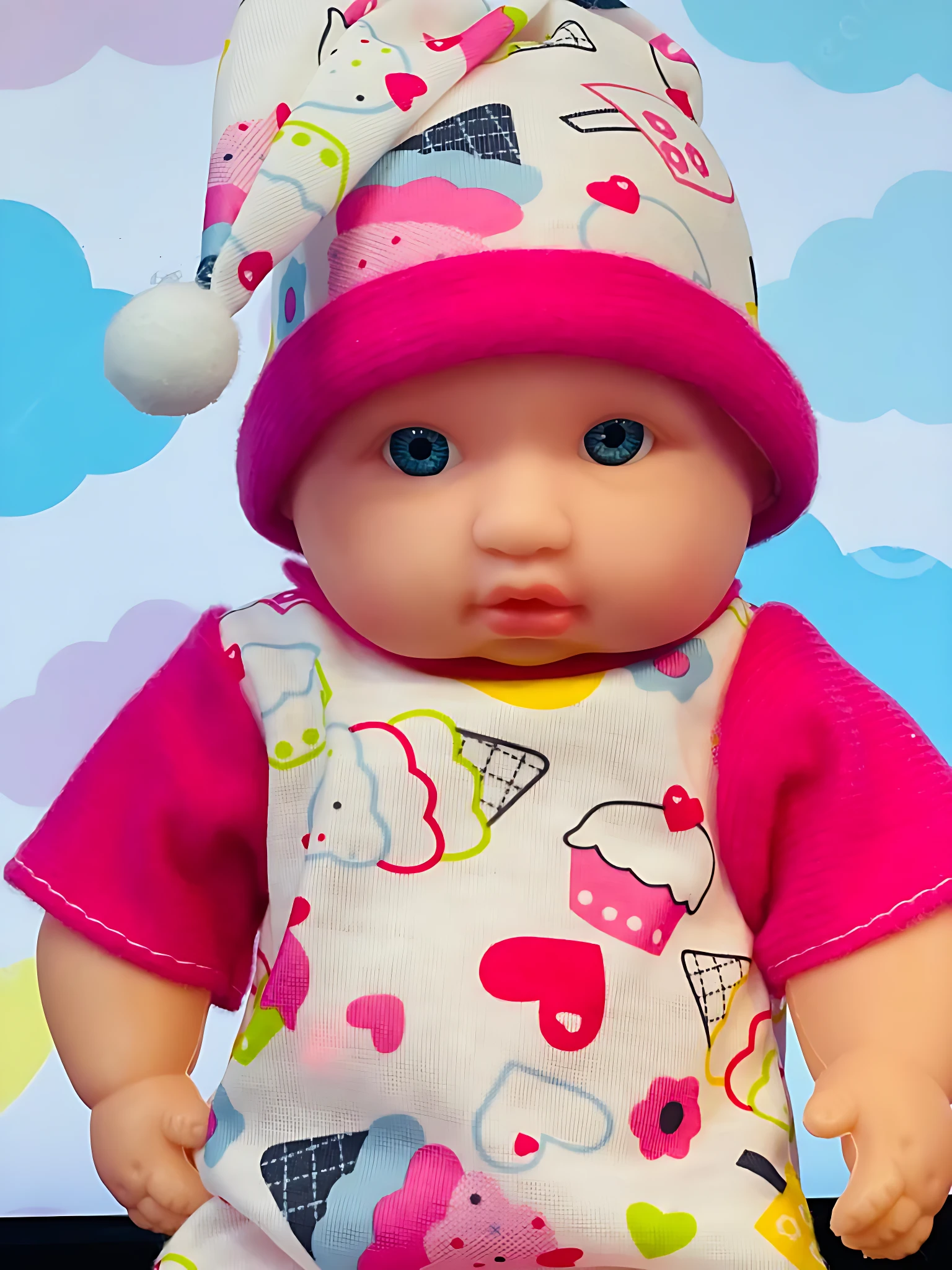 Muñeco bebé con pijama blanco y estampado de dulces, gorro rosa, ojos azules, fondo de nubes.