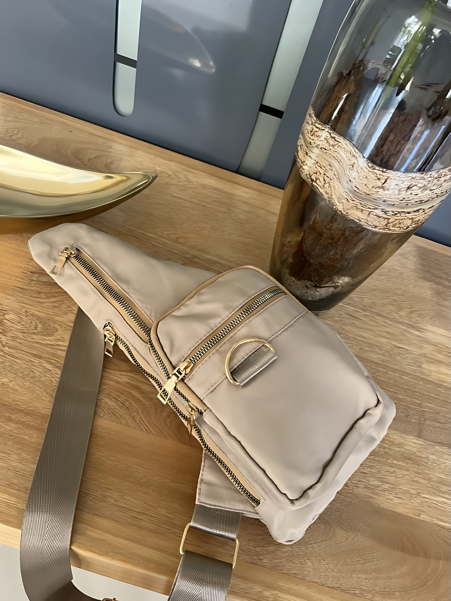Bolso bandolera impermeable color beige, con cremalleras doradas y correa ajustable, ideal para llevar lo esencial.