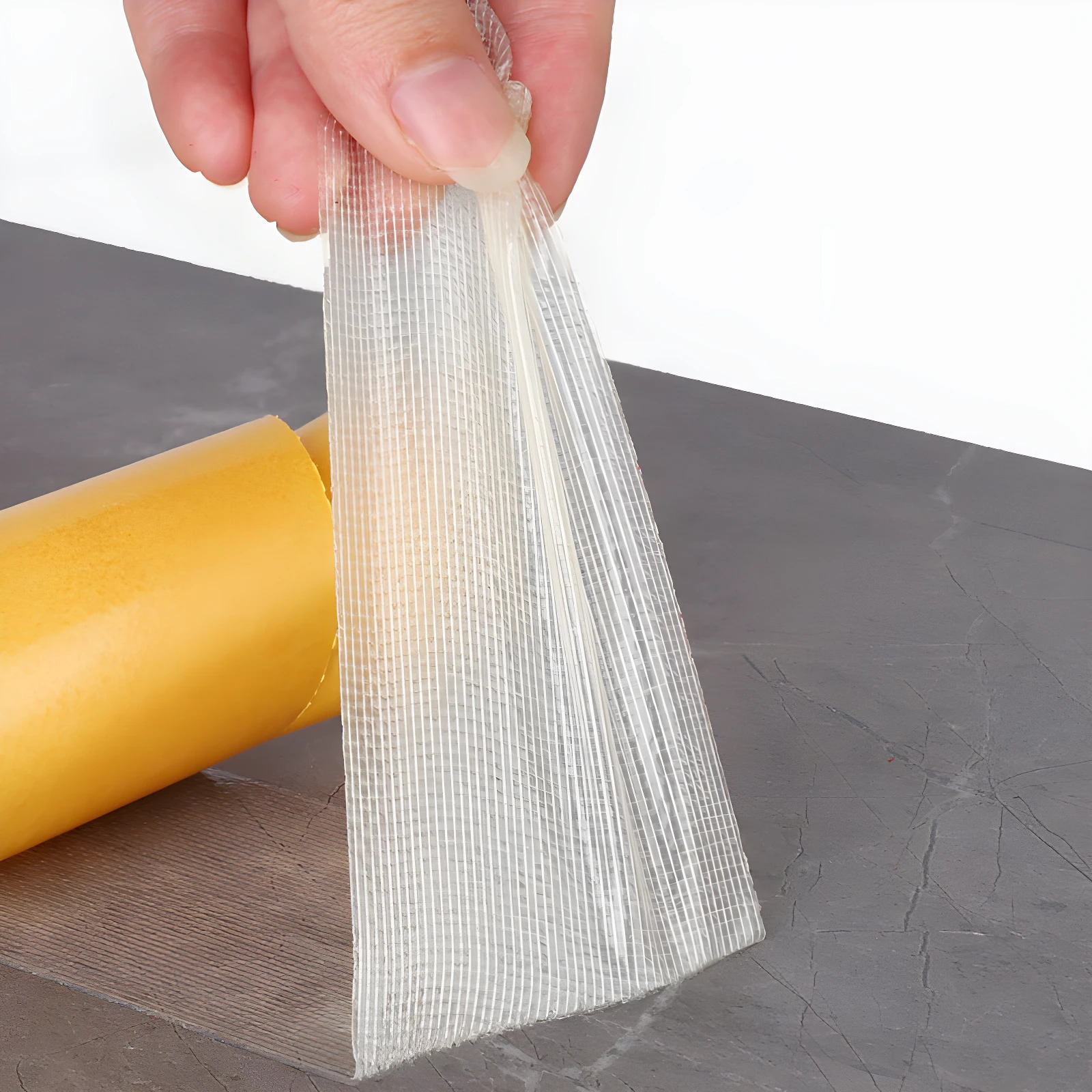 Cinta doble faz transparente con malla de refuerzo siendo retirada de un rollo amarillo, ideal para pegar objetos sin dañar superficies.