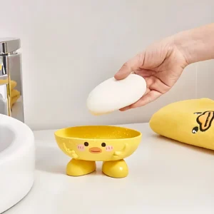 Jabonera patito amarillo sosteniendo jabón blanco, con gotas de agua, sobre superficie blanca.