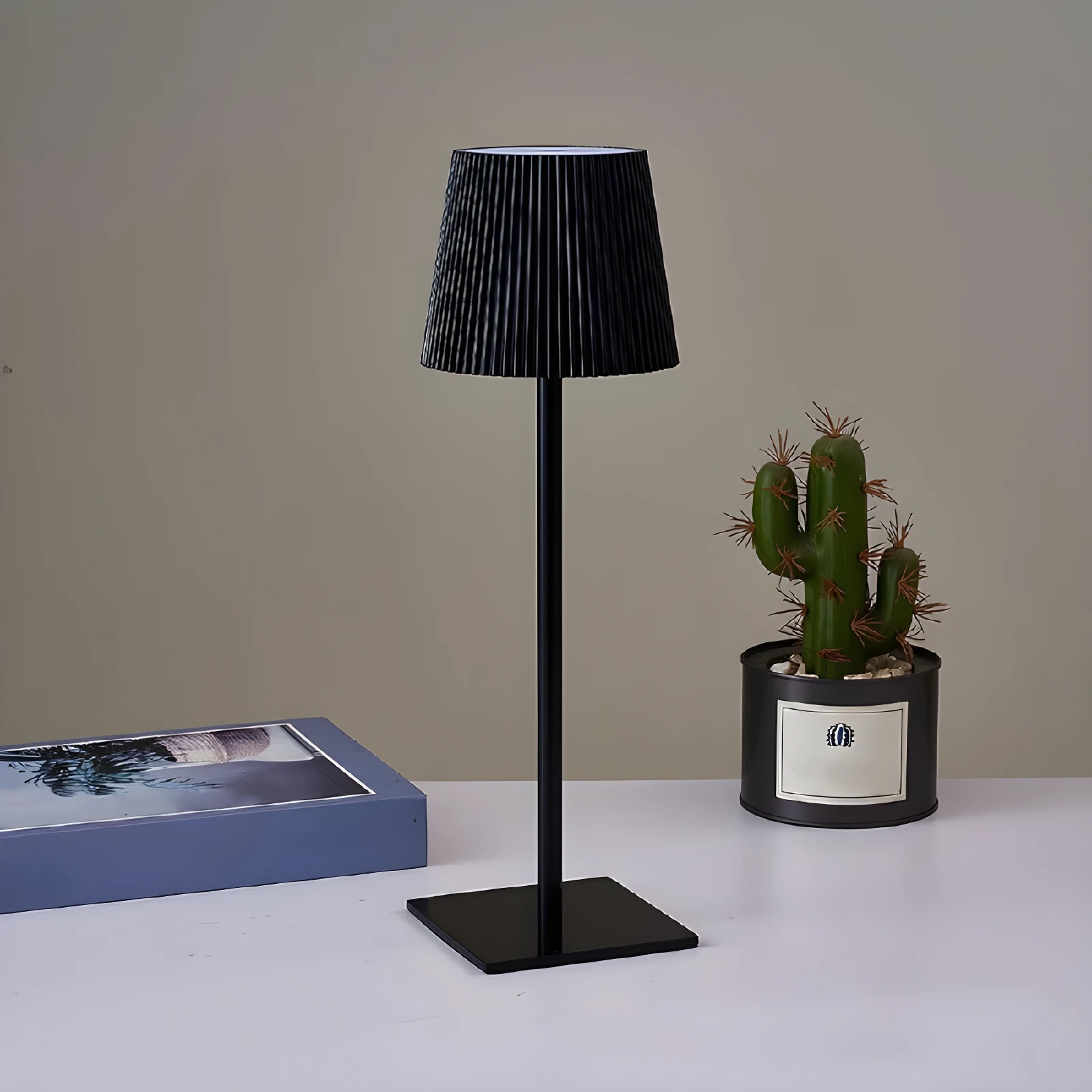 Lámpara LED negra minimalista de metal, inalámbrica, con pantalla acanalada, sobre mesa blanca junto a cactus.