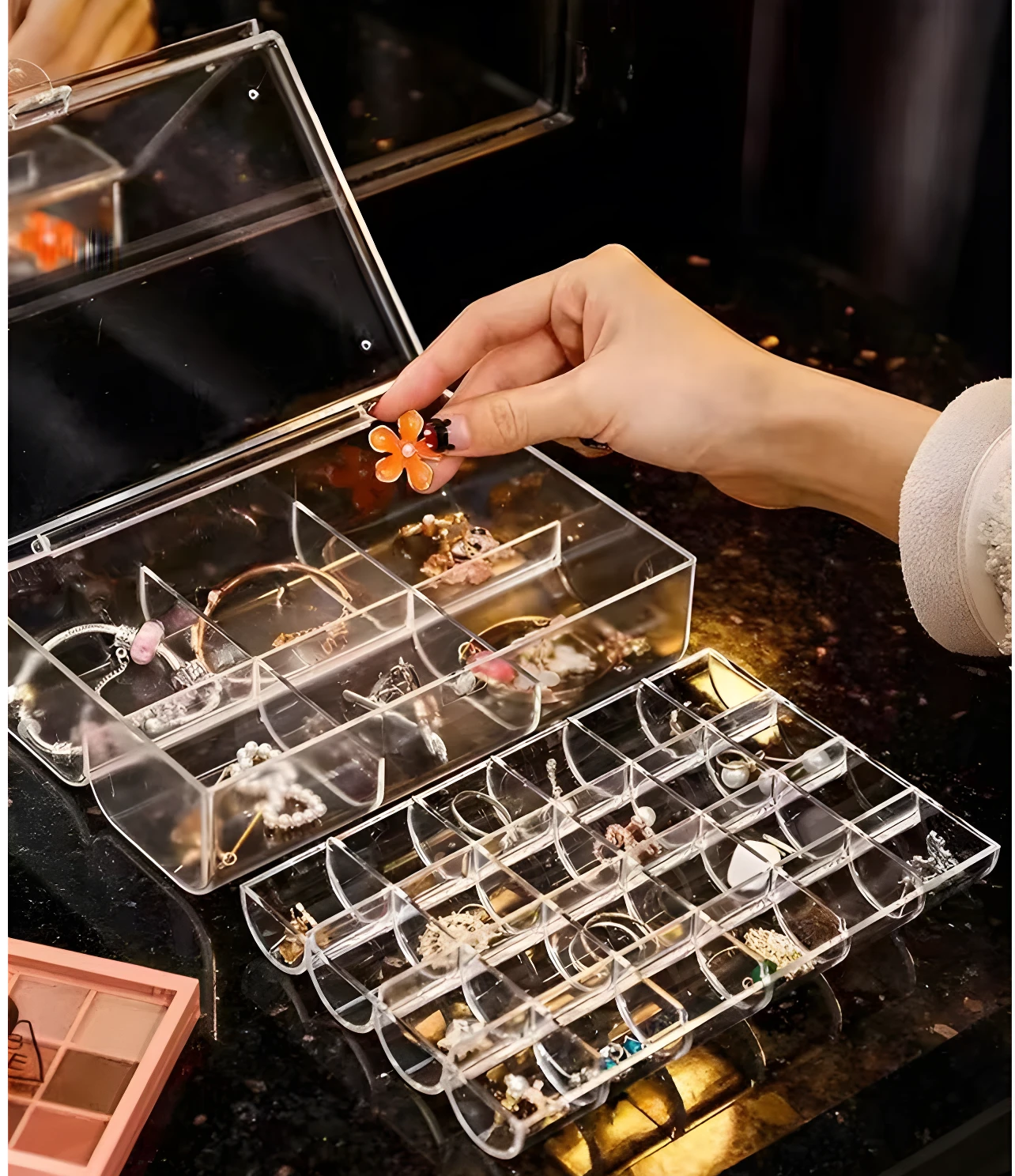Joyero apilable acrílico transparente con compartimentos organizando collares, pulseras y anillos, mano colocando detalle floral.